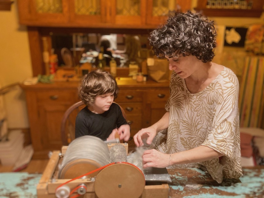 Child and adult sit at table with carding machine, wooden cabinet in background.