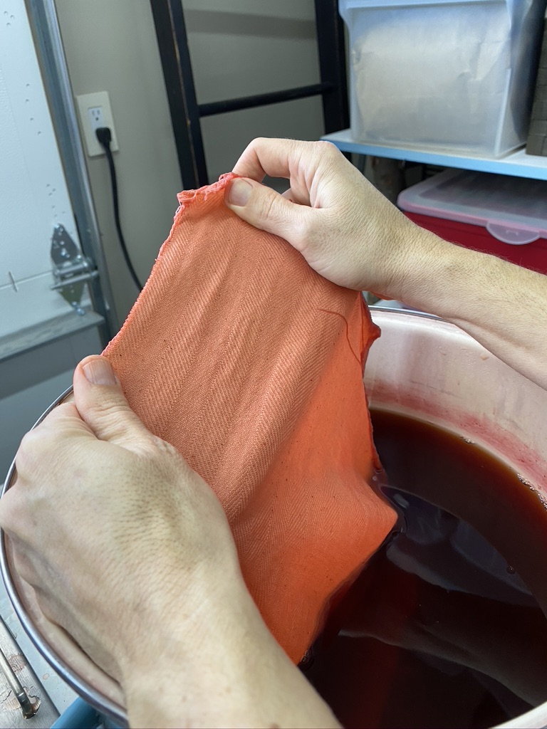 Hands stretch light orange-colored fabric above bowl with deep red liquid.
