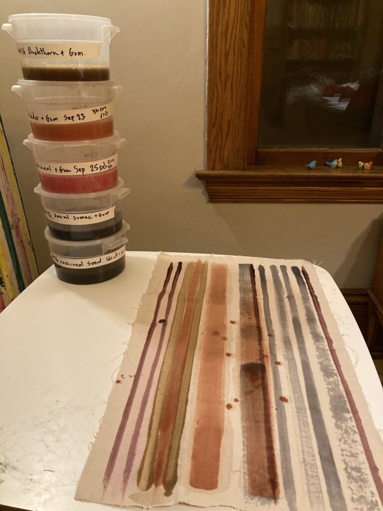 Multicolored stripes painted on fabric, which lies on white table beside stacked plastic containers of dye.