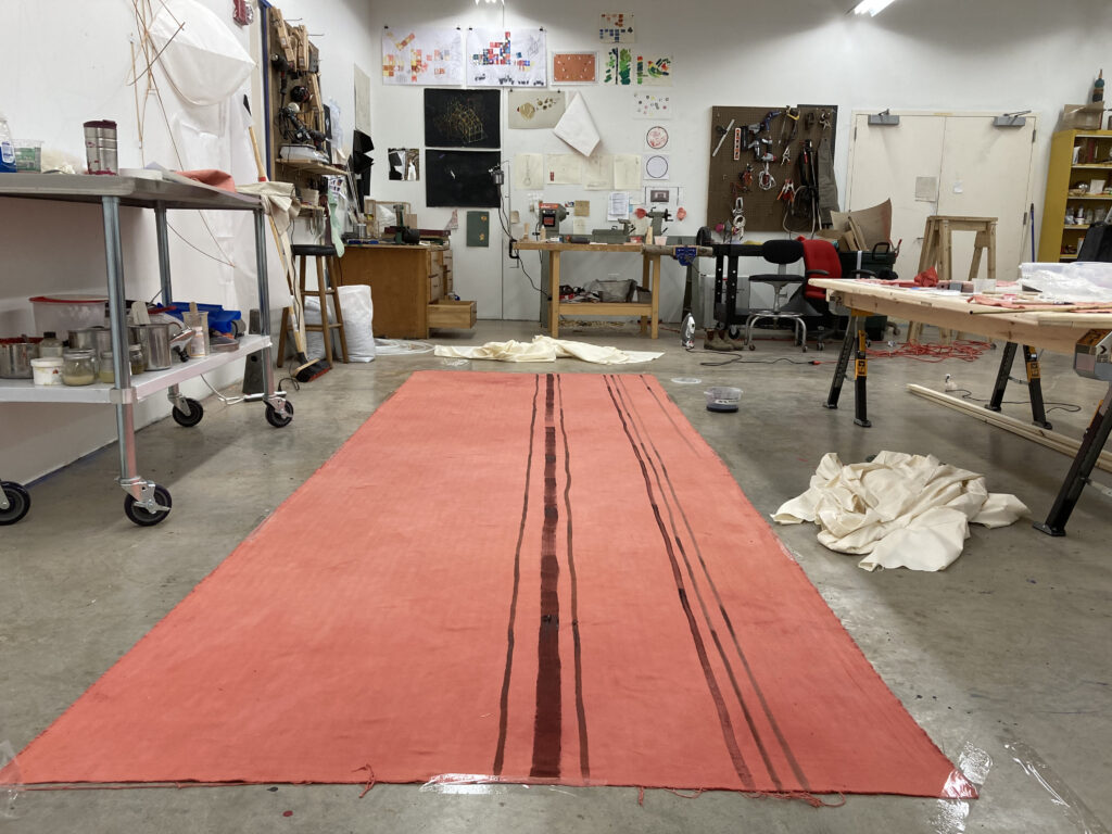 Long red fabric with stripes stretches out on floor of artist studio.