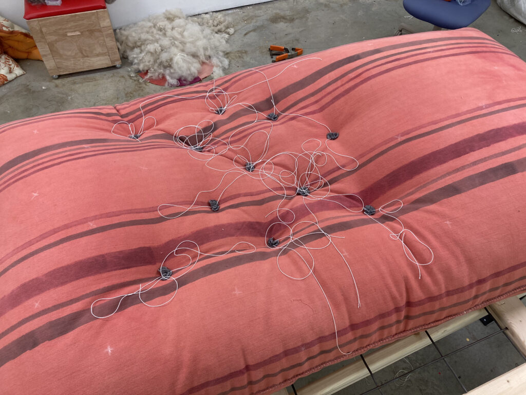 Striped red fabric mattress with scattered white threads and tufting in progress.