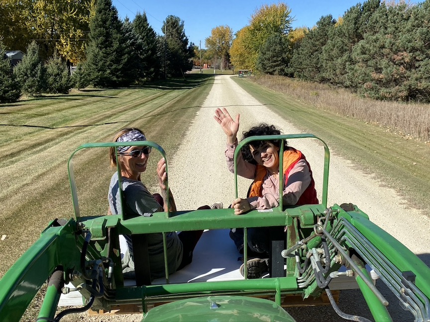 Two people turn and wave from back of green tractor, with gravel road and green grass behind them.