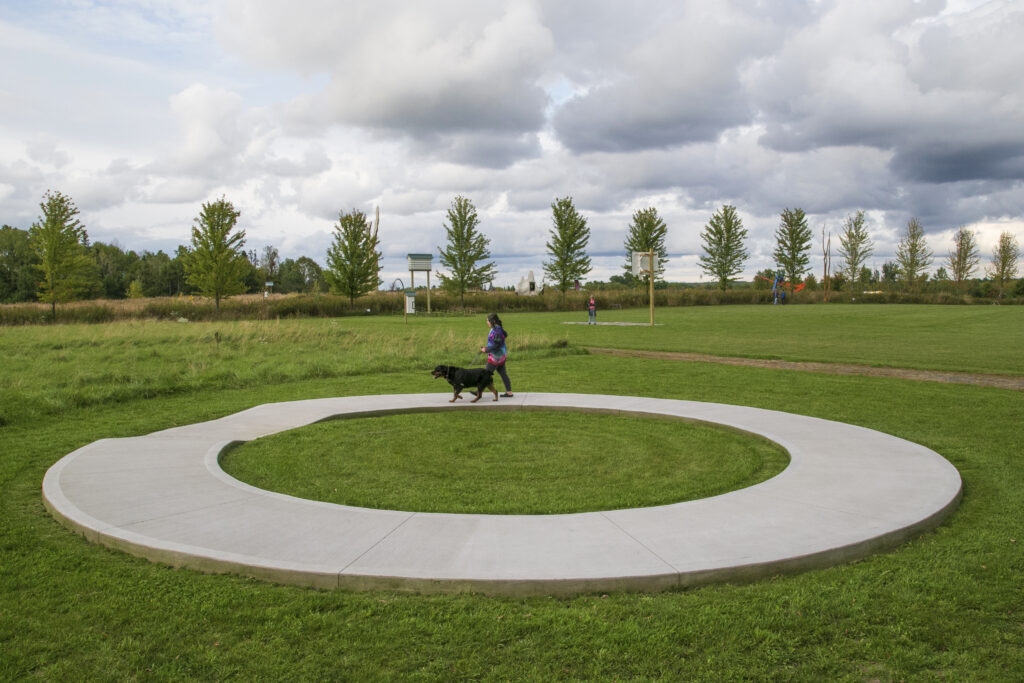 Person walks a large black dog on a sculpture shaped like circular concrete sidewalk, with open field and trees in background.