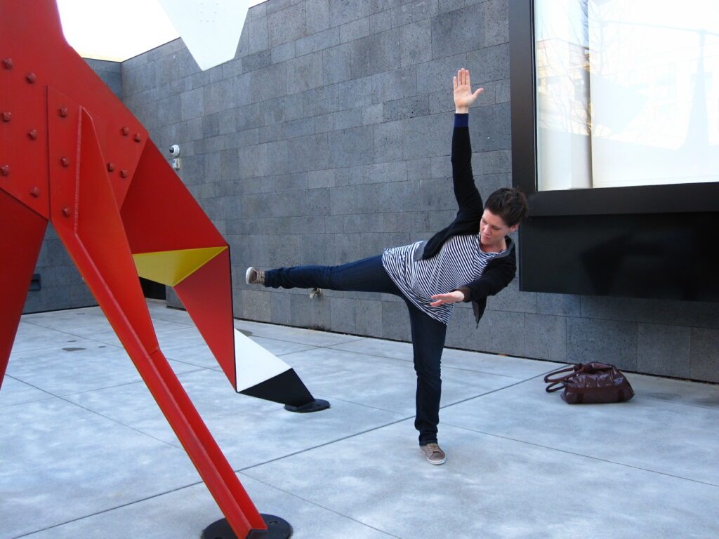 Person wearing gray jumpsuit tilts to the left side, right arm and leg raised and stretched, with red metal sculpture in foreground.