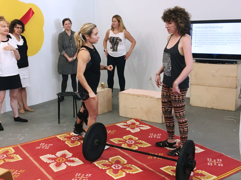 Two people stand facing each other in front of large black dumbbell on floor in gallery.