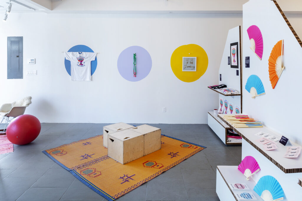 Gallery installation with orange rug and wooden boxes on floor, prints and colorful designs on wall.
