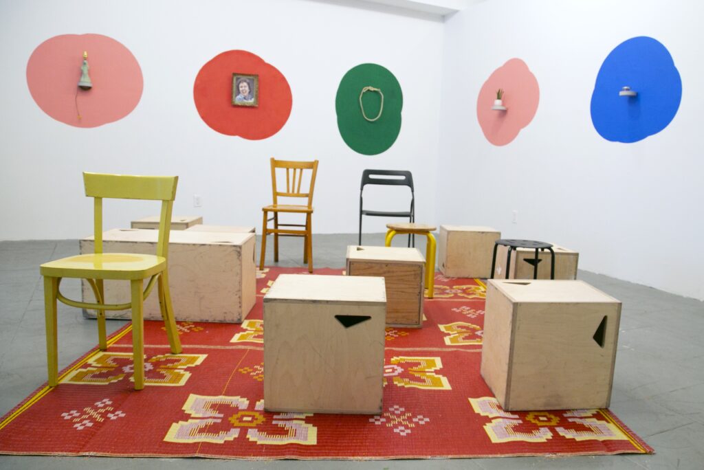 Gallery with orange rug, wooden boxes, various chairs, and multicolored designs on white wall. 