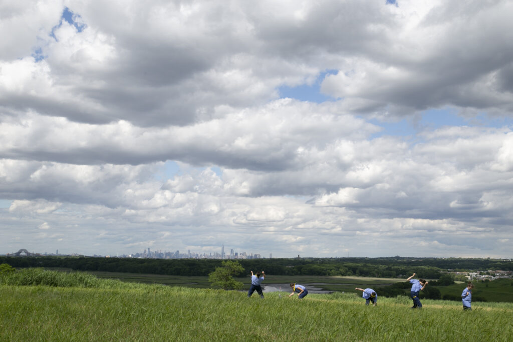Group of people wearing blue dances in open, grassy field, with large blue sky with white clouds.