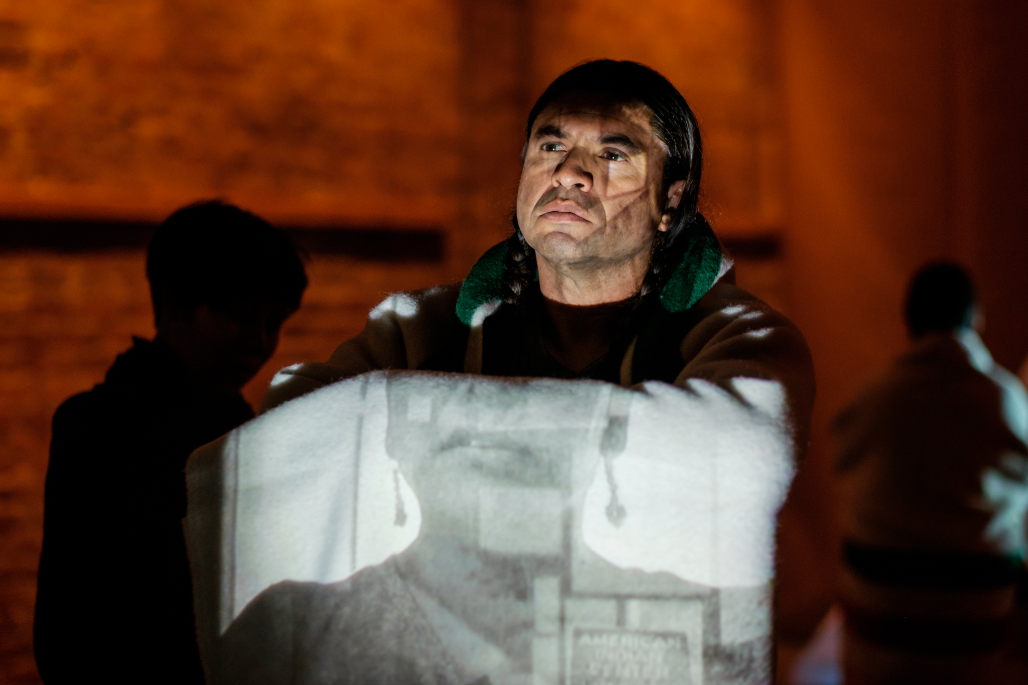 Person with long, dark hair pulled back crosses his arms in front of his body, draping fabric on which a black and white image of a face is projected, with warm lighting in background.