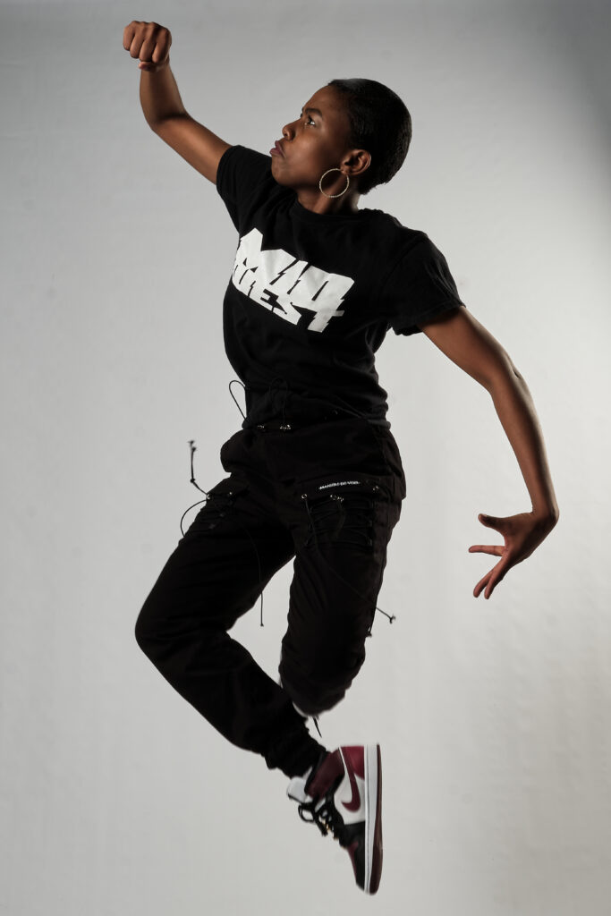 Person with dark skin and short hair, wearing black sweatpants, t-shirt, and sneakers, jumps in the air with their fist raised.