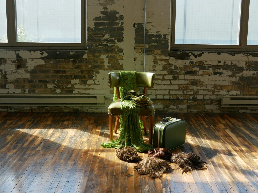 Green chair draped with fabric sits is surrounded by suitcase and many wigs on wooden floor, with sunlight streaming in window.