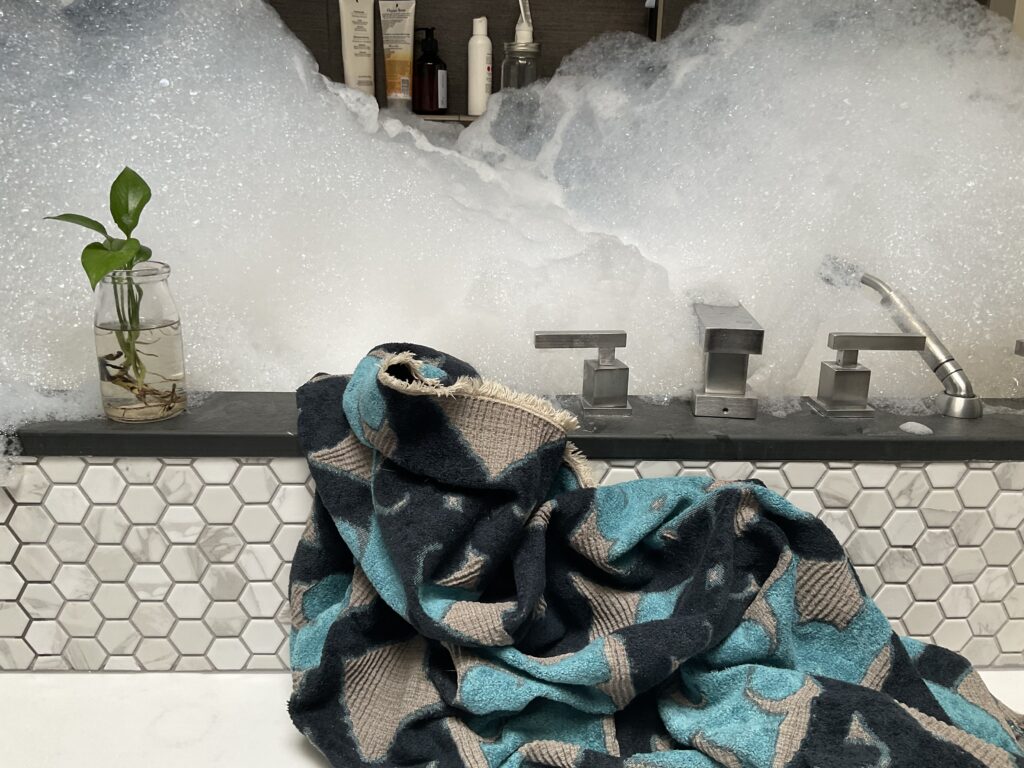 Blue patterned blanket approaches a bathtub overflowing with bubbles.