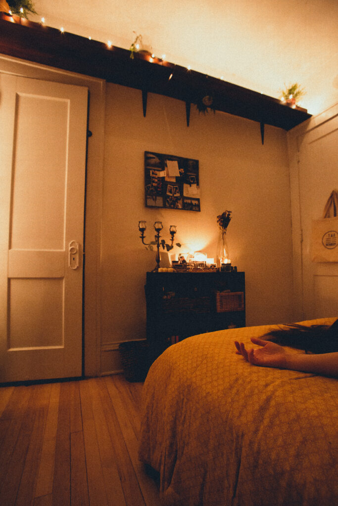 A warmly lit photo of a bedroom. A hand and long dark hair is visible resting on the corner of the bed, with the rest of the body out of frame.