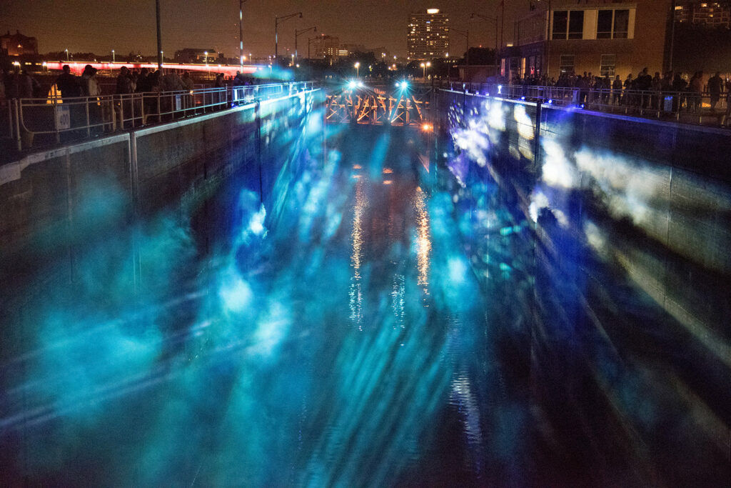 Beams of blue and purple light illuminate fog in a lock, with people standing at upper rim.