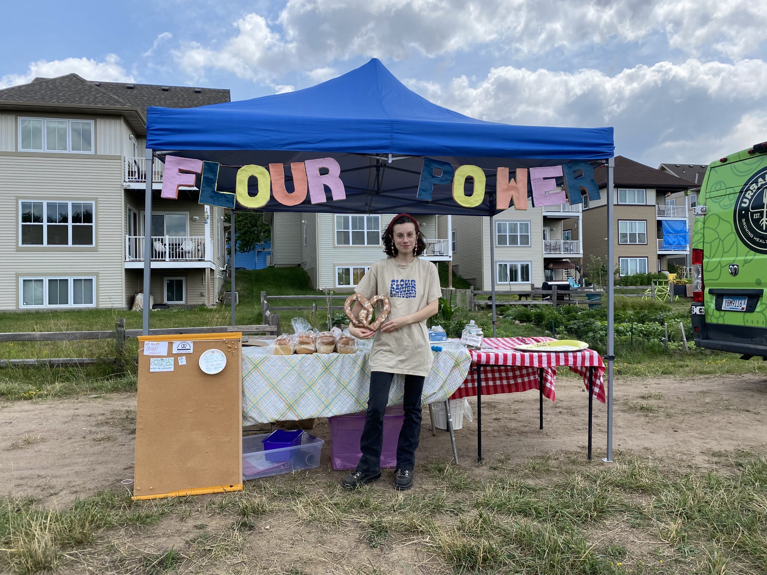 Person holding large soft pretzel stands in front of booth with baked goods, blue awning, and large multicolored letters reading FLOUR POWER.