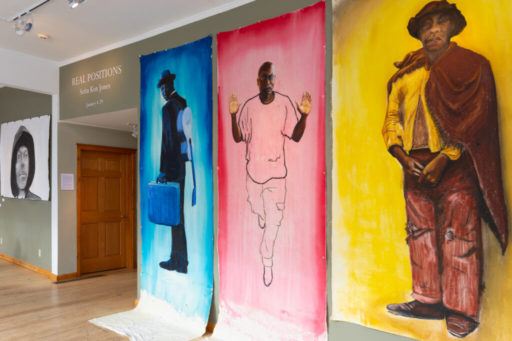 Three large, unframed canvases hang on wall and cascade down to the floor. Each contains a full-length figure of a Black man, with blue, red, and yellow backgrounds from left to right.