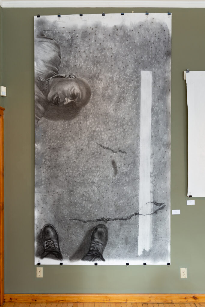 Painting in grayscale hangs on beige wall. Painting depicts Black man with eyes closed on the ground, with boots in bottom of frame.