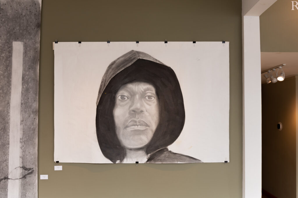 Painting in grayscale hangs on wall, depicting Black man in dark hoodie looking directly at viewer.