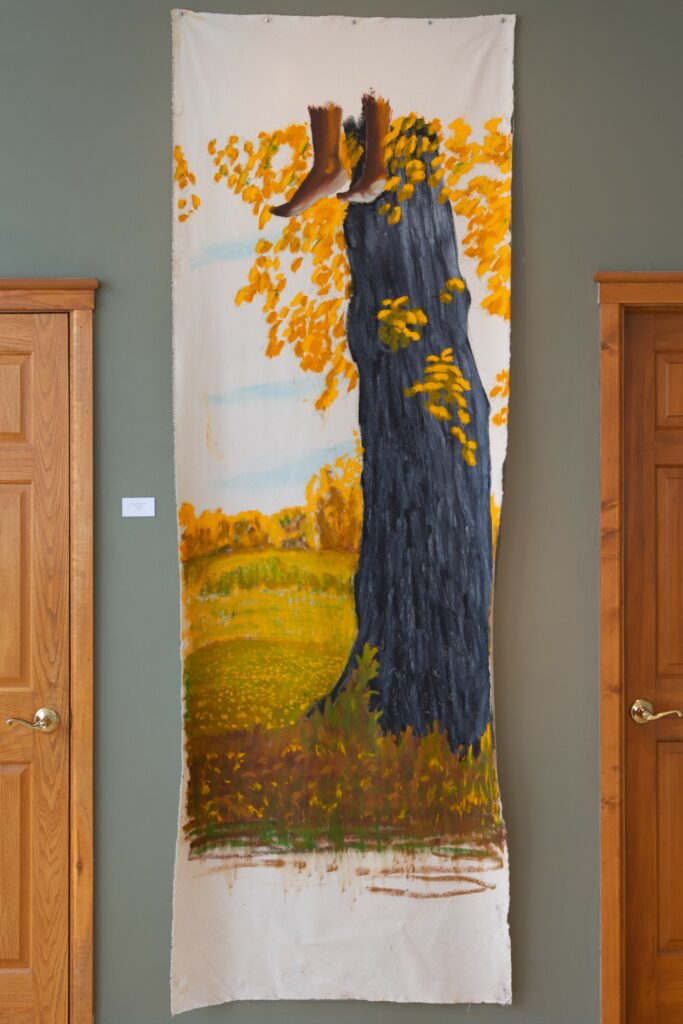 Tall painting hangs on wall, depicting tree trunk with yellow leaves, and feet with dark skin hanging at top of frame.