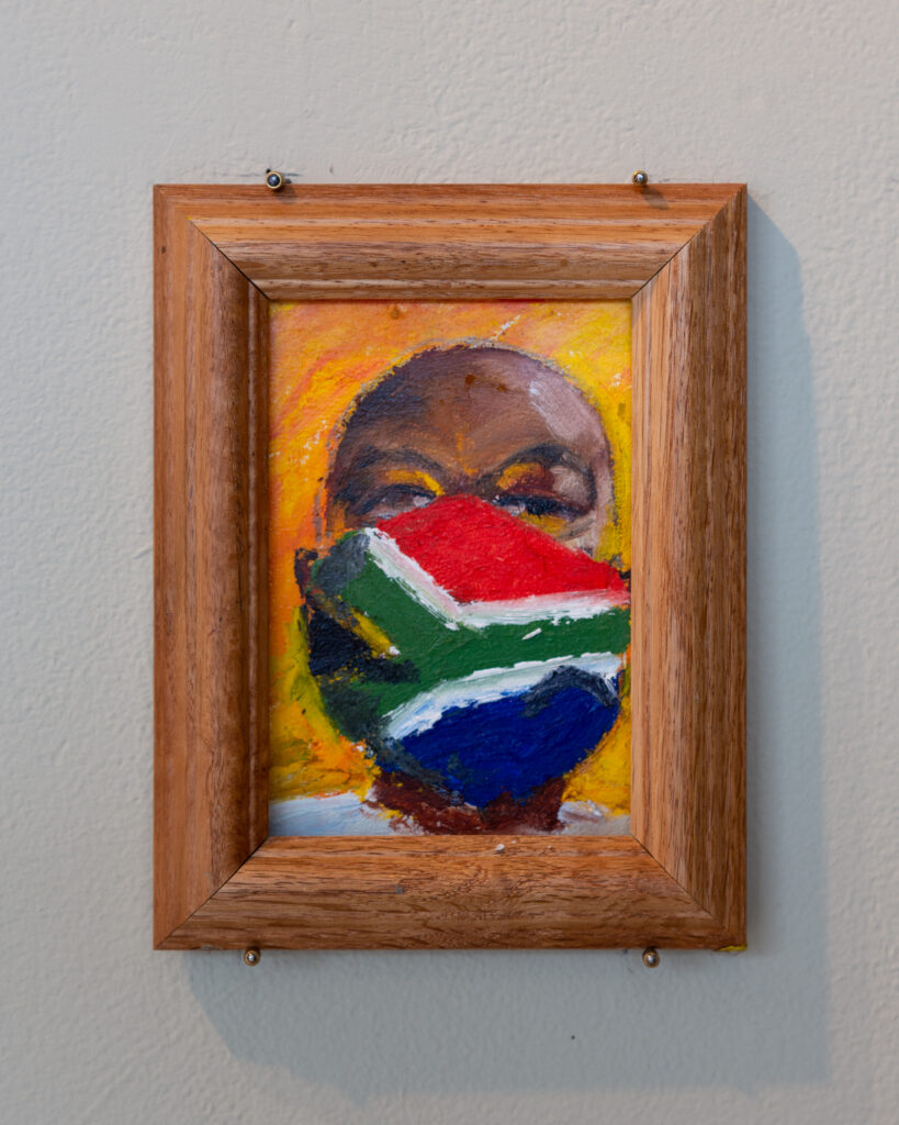 Painting with thick wooden frame hangs on wall, depicting a face with dark skin wearing a red, green, blue, and white flag mask.
