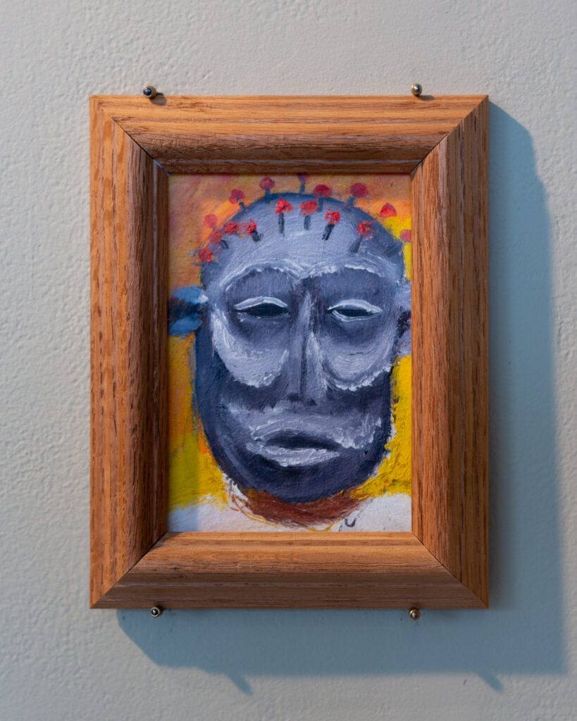 Painting with thick wooden frame hangs on wall, depicting angular gray mask with red pins on head.