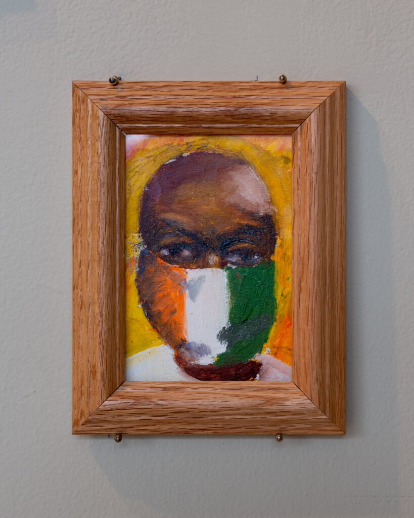 Painting with thick wooden frame hangs on wall, depicting a face with dark skin wearing an orange, white, and green flag mask.