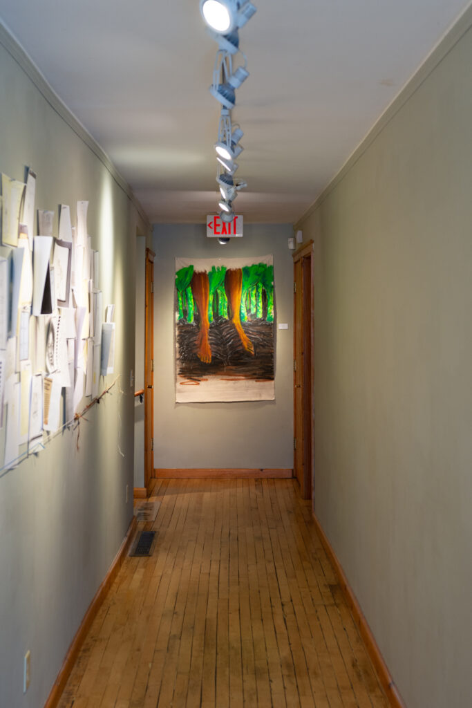 Hallway with beige walls and light wood floor, with drawings and collage on left wall and painting of rooted feet with dark skin on back wall.
