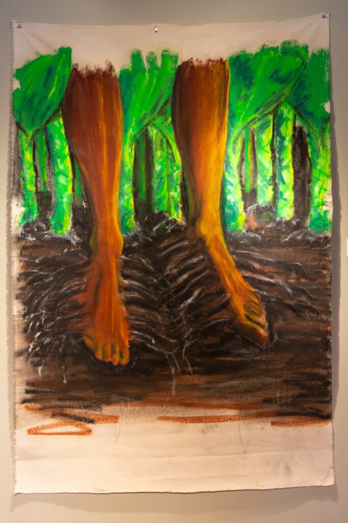 Painting hangs on wall, depicting feet with dark skin sprouting roots into dark brown soil, surrounded by green stalks.