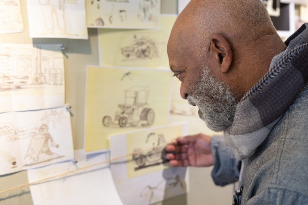 Person with dark skin and gray beard looks down and touches drawings pinned on wall.