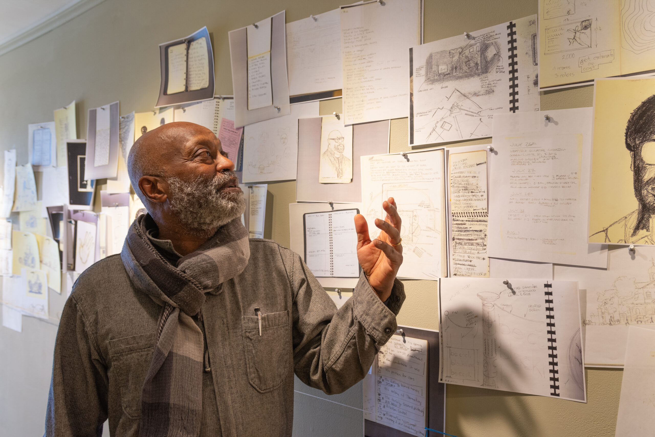 Person with dark skin, gray beard, gray shirt and scarf gestures with open hand while looking up at beige wall covered with many small drawings.