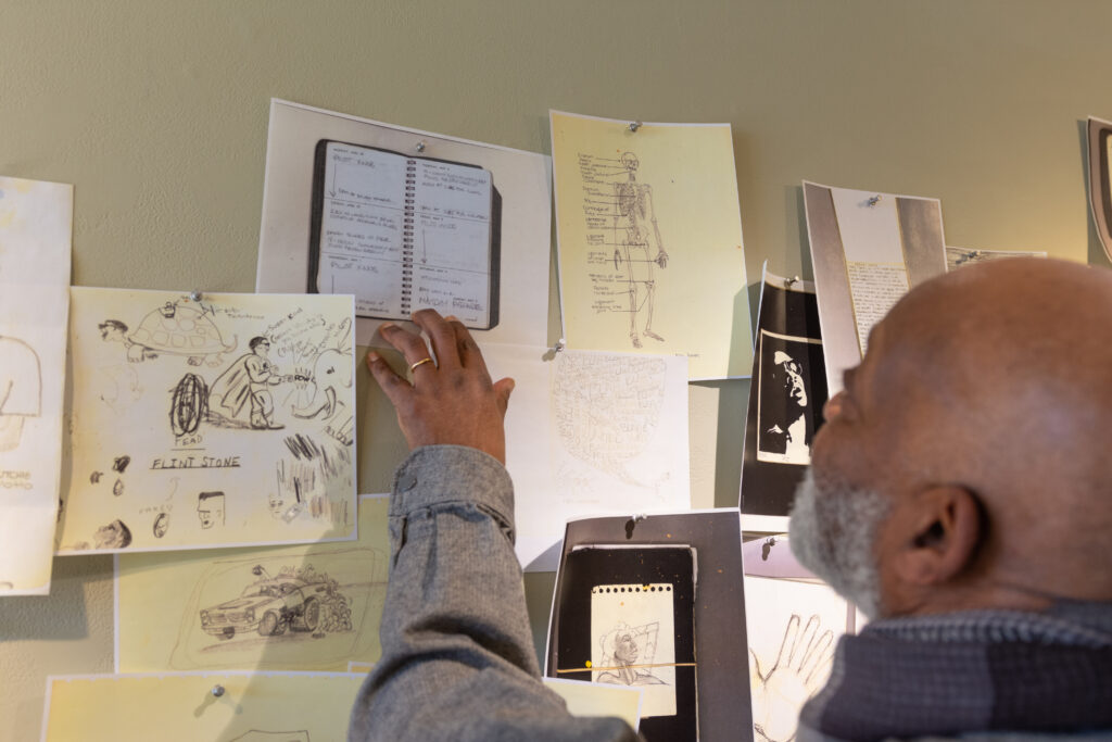 Person with dark skin and gray beard reaches up to touch drawings and collages on wall.