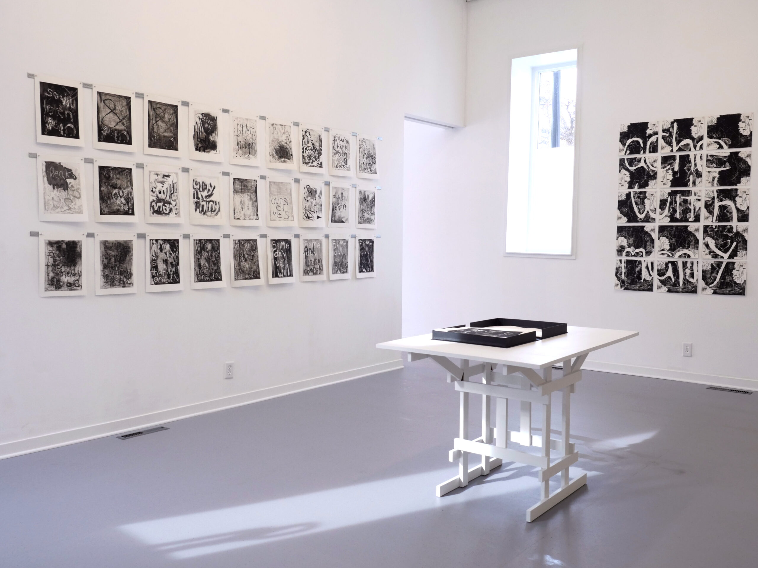 The interior of Dreamsong’s gallery, which has white walls and a flat gray floor. This image, taken from the corner of the room, shows prints hanging on two adjacent walls, and a white table in the center of the room. On top of the white table is a short black box, opened, with prints stacked inside.