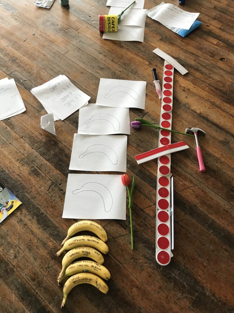 Line of yellow bananas, white papers with banana tracings, red reflectors, pink hammer, and other items on brown wood floor.