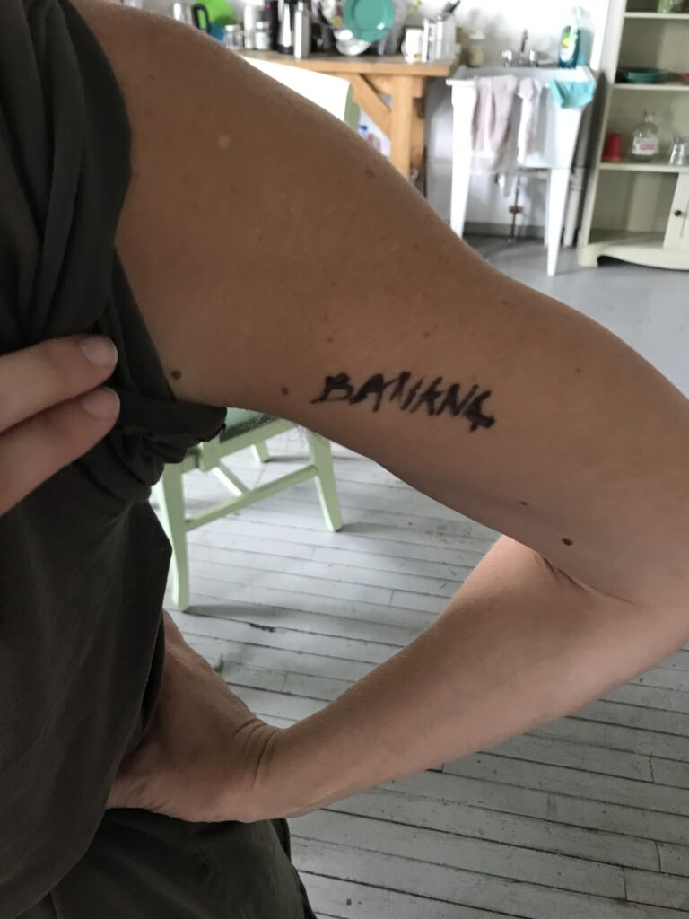 Person with light skin holds their hand to their hip, with BANANA written in black sharpie on their tricep.