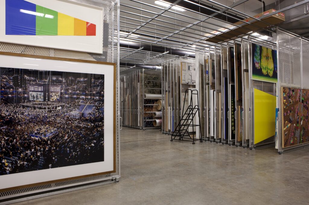 Art storage, with many framed 2D pieces hung in dense racks, with ladder, concrete floor, and fluorescent lighting.