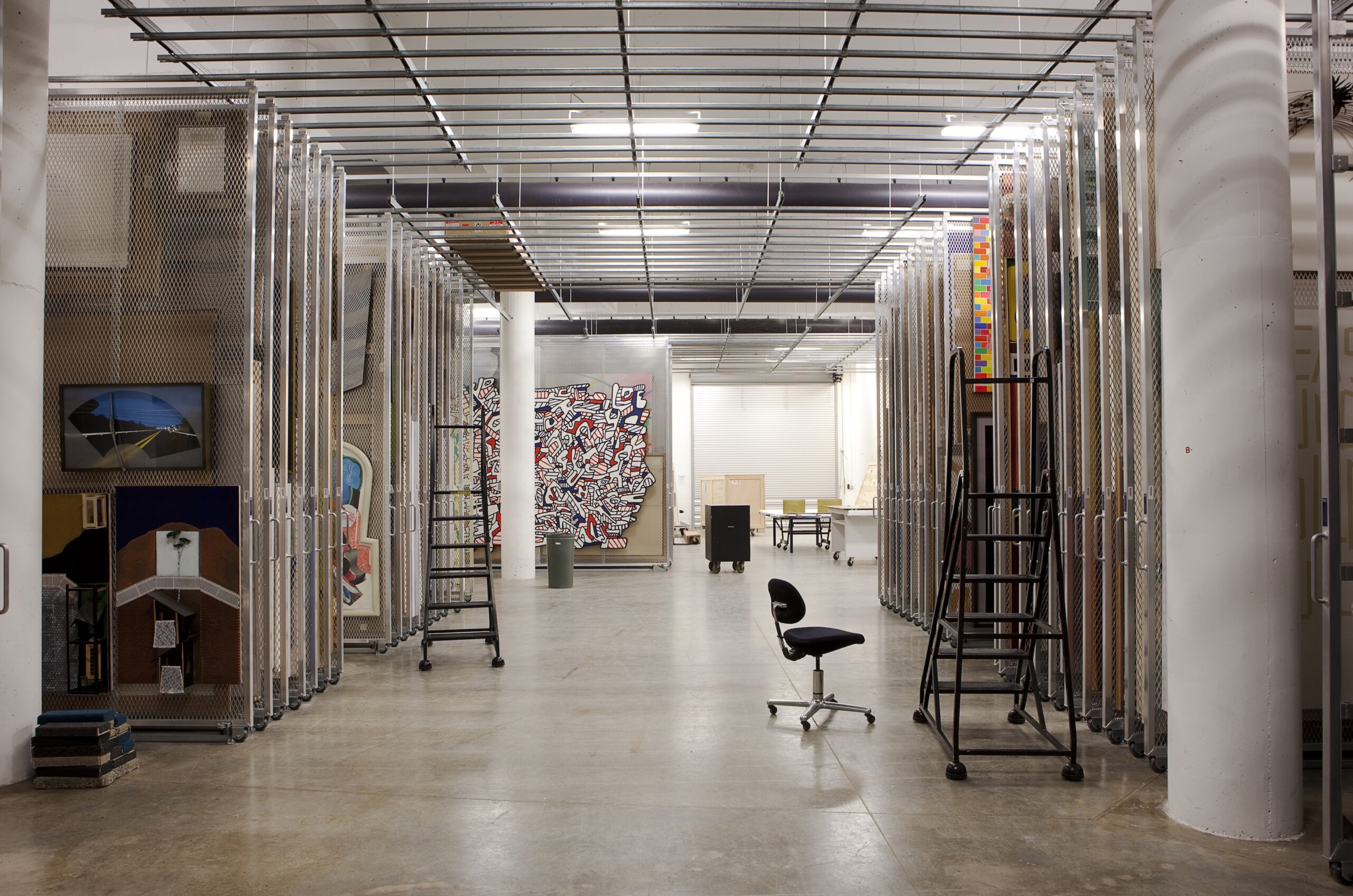 Art storage, with many framed 2D pieces hung in dense racks, with chair, ladder, concrete floor, and fluorescent lighting.