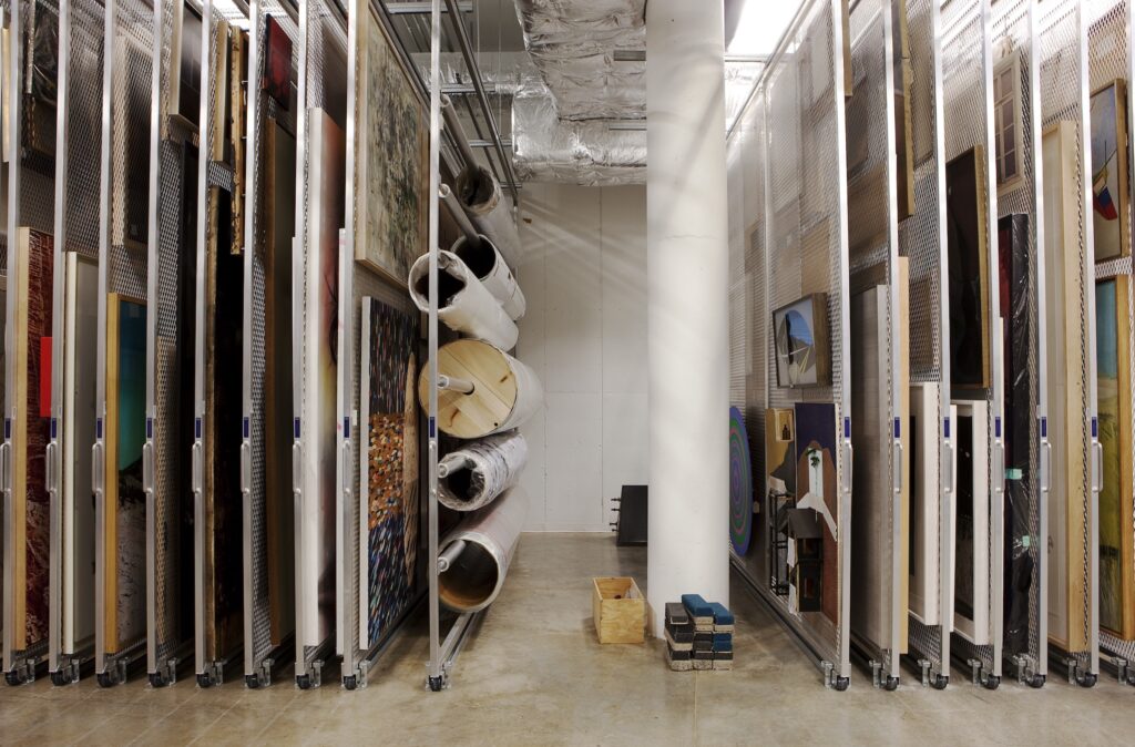 Art storage, with many 2D and rolled artworks hung on racks, with white column and concrete floor.