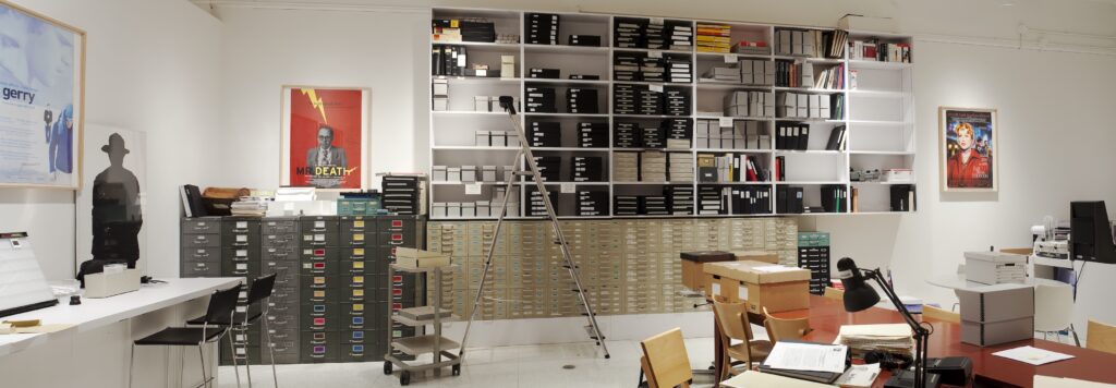 Room with gray and beige file cabinets, tall shelving, filing boxes, ladder, carts, tables, and chairs.