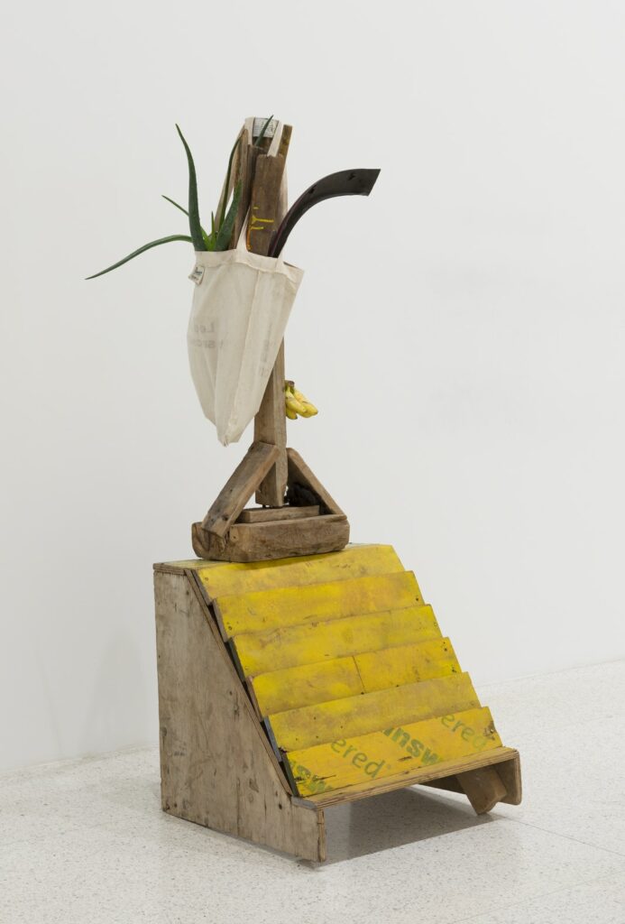 Sculpture with wooden base with yellow stairs, wooden stand, bag with green plant fronds, and hanging bunch of bananas.