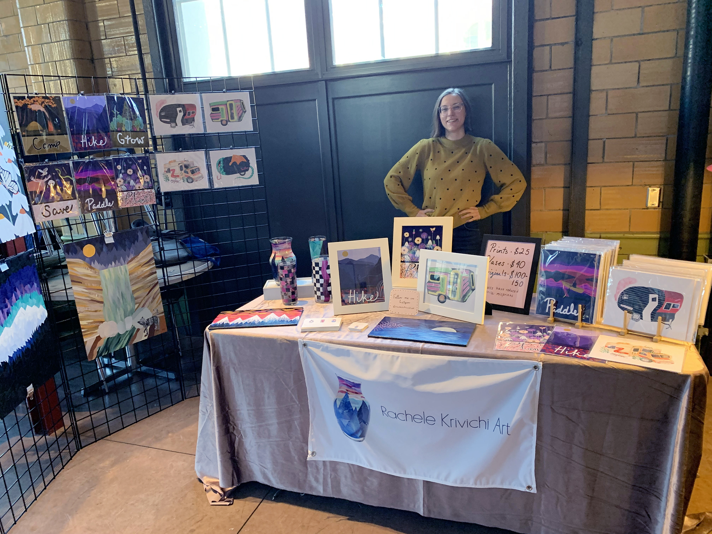 Person stands with hands on hips behind a table with artwork on display.