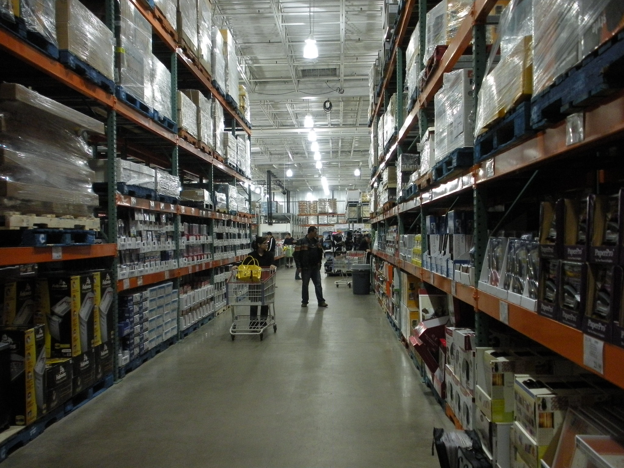 Two people and shopping cart stand at the end of a long aisle of big box store with tall orange shelving.