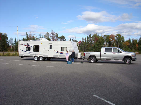 Silver pickup truck with white RV in large, empty parking lot and blue sky.