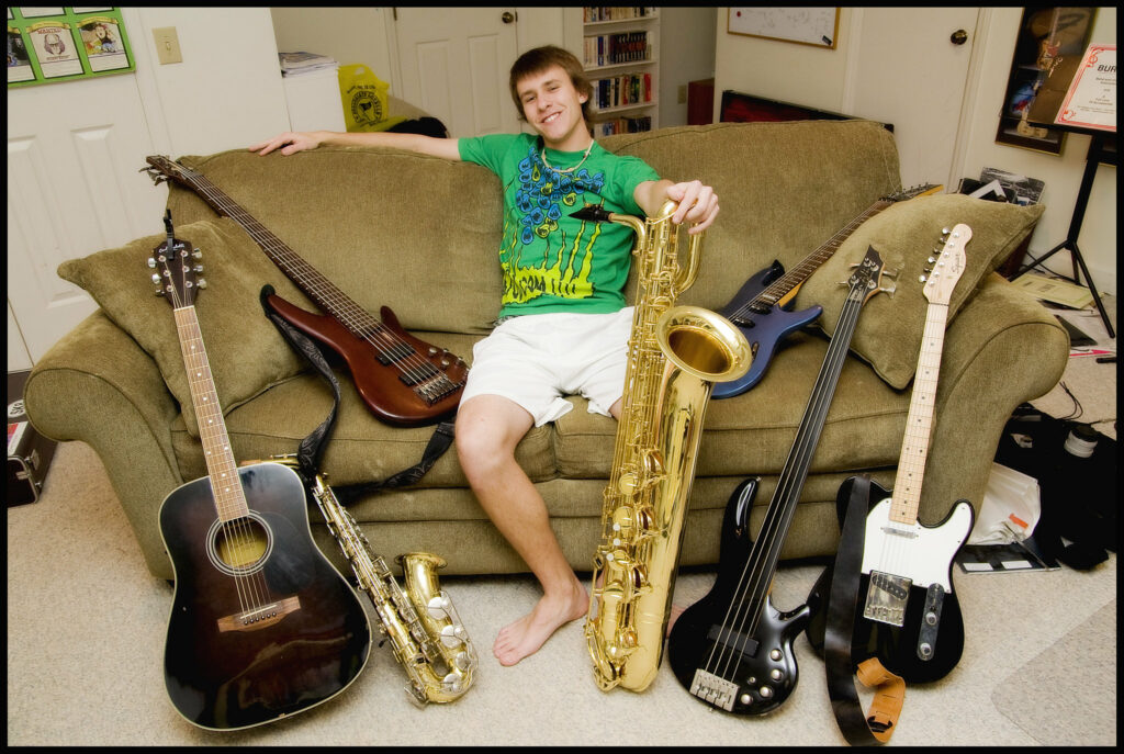Person with khaki shorts, green t-shirt, and light skin sits on beige couch surrounded by many musical instruments, including acoustic and electric guitars, basses, and two saxophones.