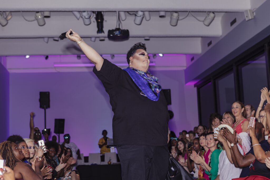 Person with contoured makeup strikes pose with their arm in the air, with runway and crowd behind them.