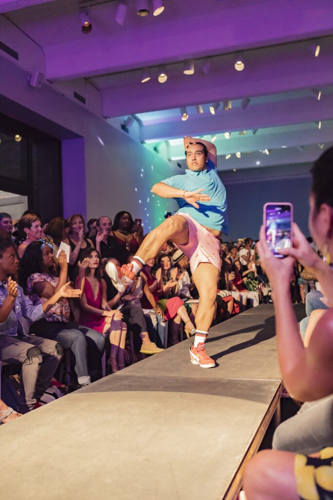 Person strikes a pose on end of runway with leg in the air and crowd on either side.