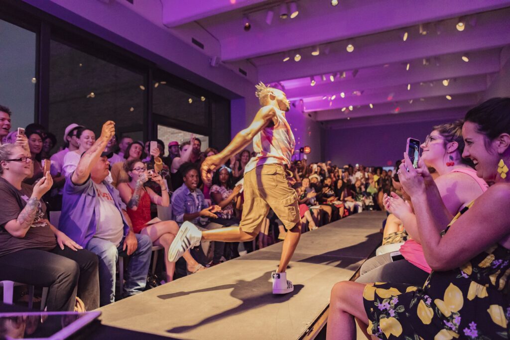 Person with dark skin and shorts lands a jump away from the camera, with runway, crowd, and purple lighting in background.