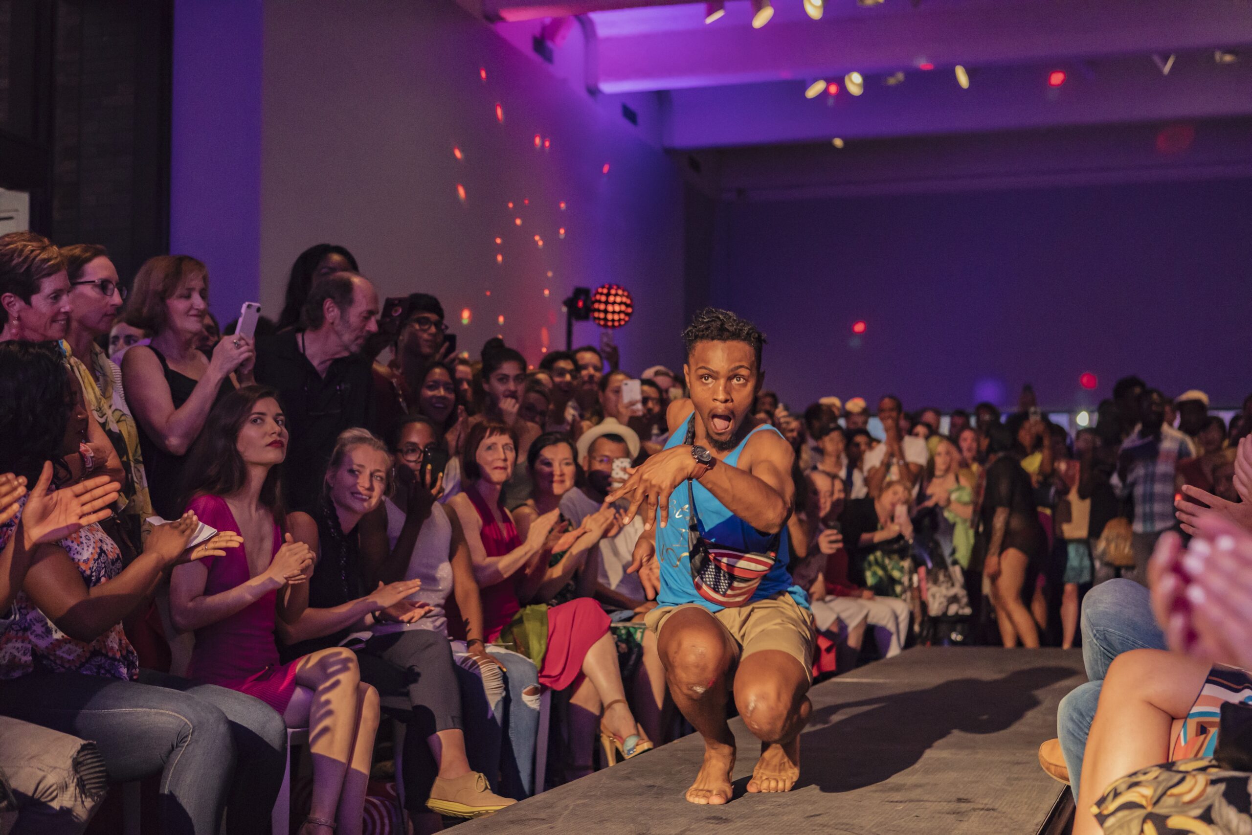 Person with dark skin performs duck walk on runway, with crowd and purple lighting behind them.