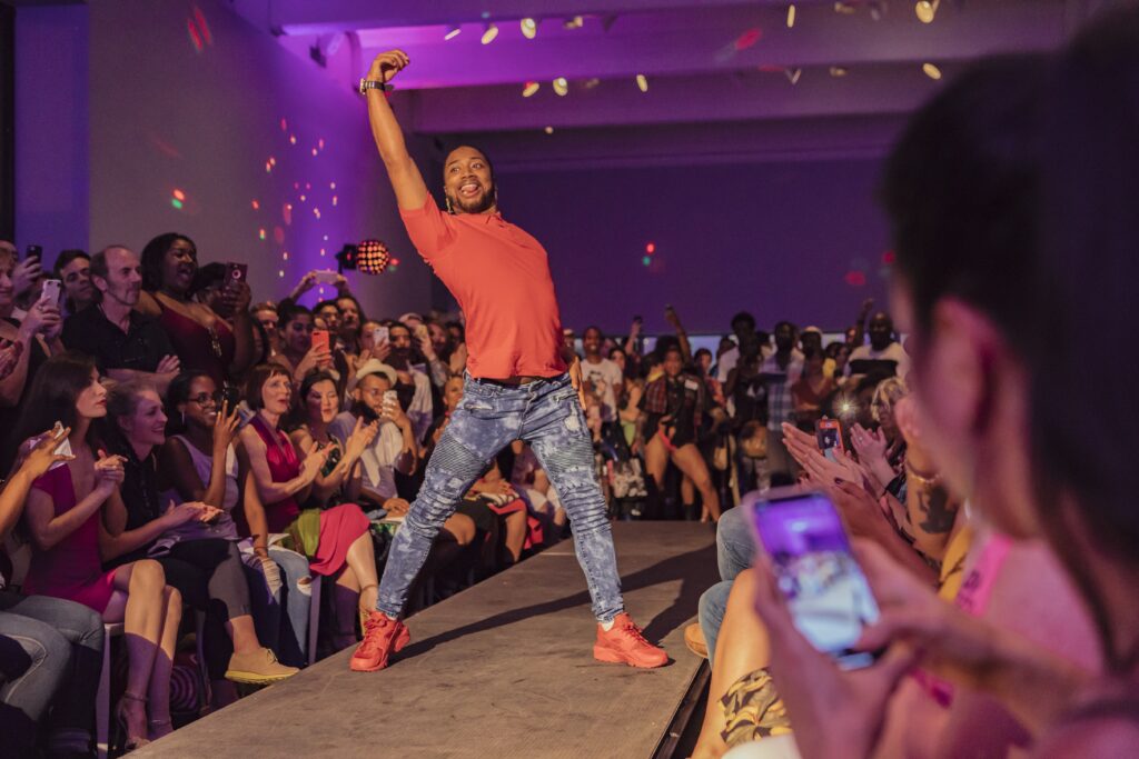 Person with dark skin, jeans and red sneakers strikes a pose with arm in the air on end of runway, with crowd behind them.
