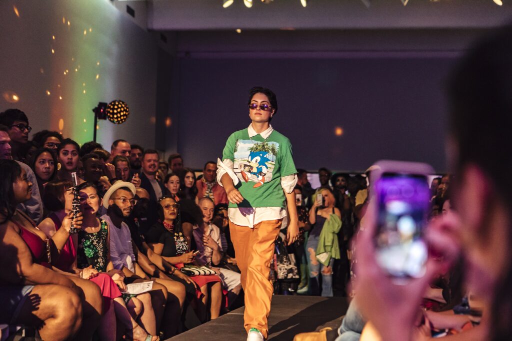 Person wearing sunglasses walks a runway with crowd on either side of them.