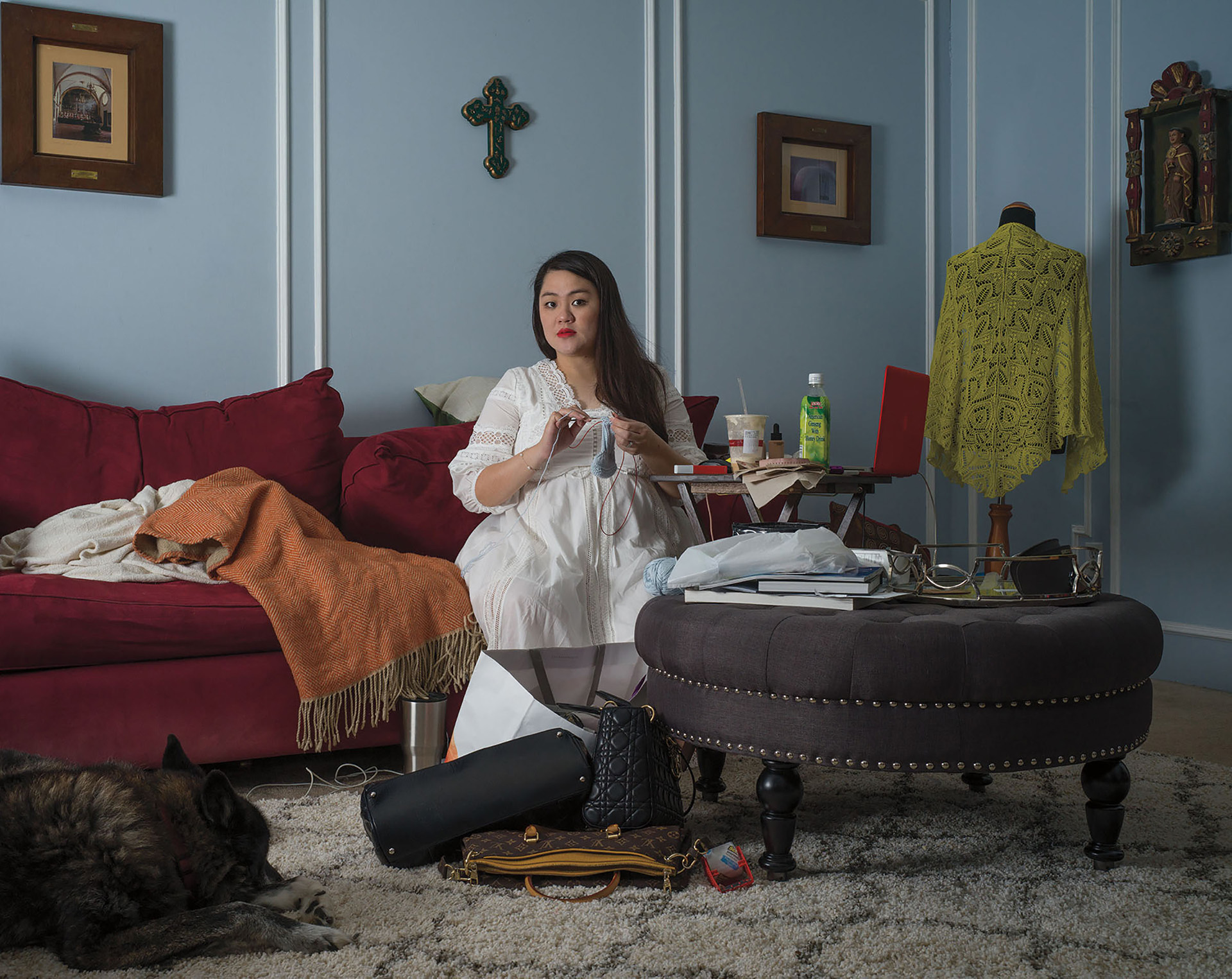 Woman with long dark hair, white dress, and red lipstick knitting on a red couch in a living room.